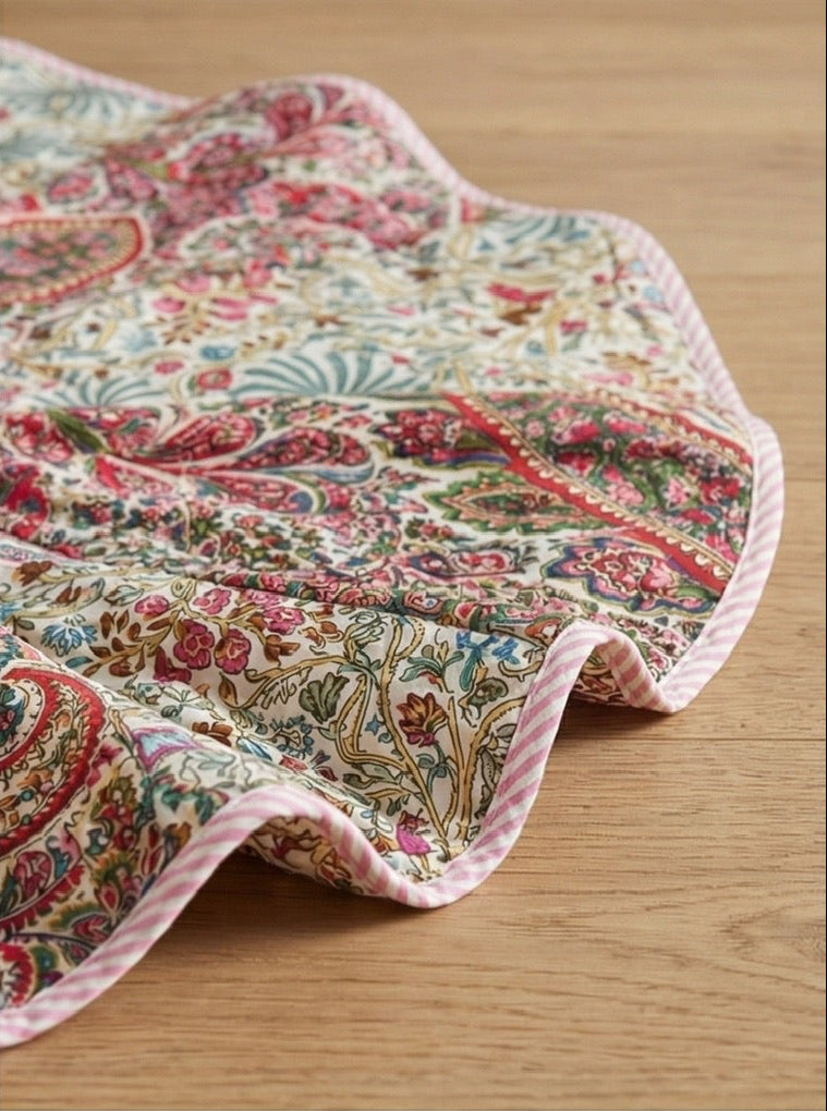 Patterned Christmas tree skirt and tablecloth on a wooden floor with a decorated tree and fireplace.
