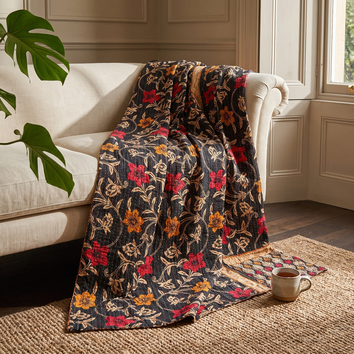 The Meadow Journey Vintage Cotton Kantha Throw displays a vibrant floral pattern with red and orange blossoms, tan vines, and visible kantha stitching on a black background, with a decorative gold patterned border.
