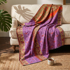 A Loom Passage Vintage Cotton Kantha Throw in shades of purple, gold, orange, and red features intricate floral, geometric, and striped patterns, draped elegantly over a beige sofa.
