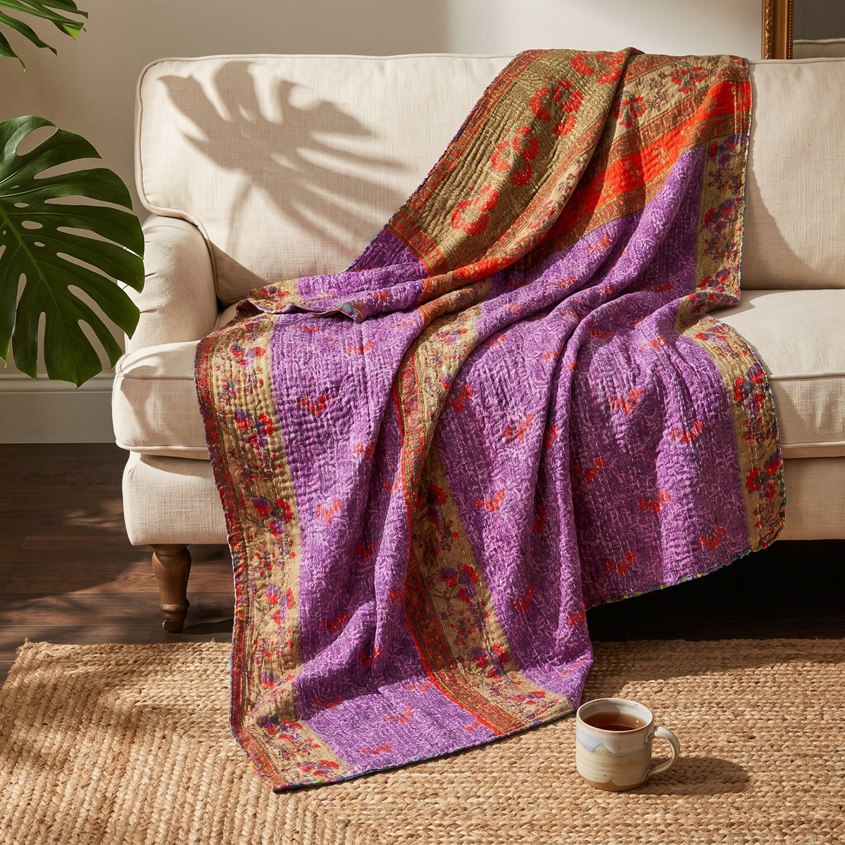 A Loom Passage Vintage Cotton Kantha Throw in shades of purple, gold, orange, and red features intricate floral, geometric, and striped patterns, draped elegantly over a beige sofa.
