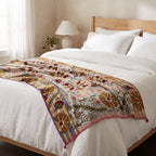 A multicolor patterned throw blanket with floral and geometric motifs is draped across a neatly made bed with white linens, a wooden headboard, and a bedside lamp in a softly lit bedroom.